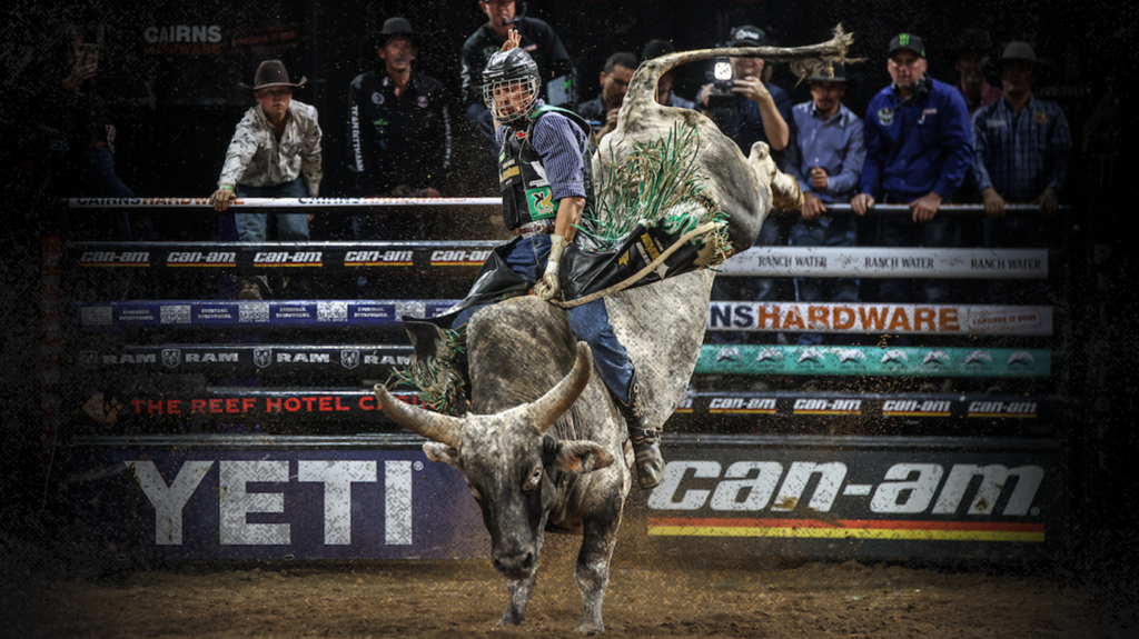 A man in a cowboy hat rides a bucking bull in a rodeo arena, surrounded by cheering spectators.